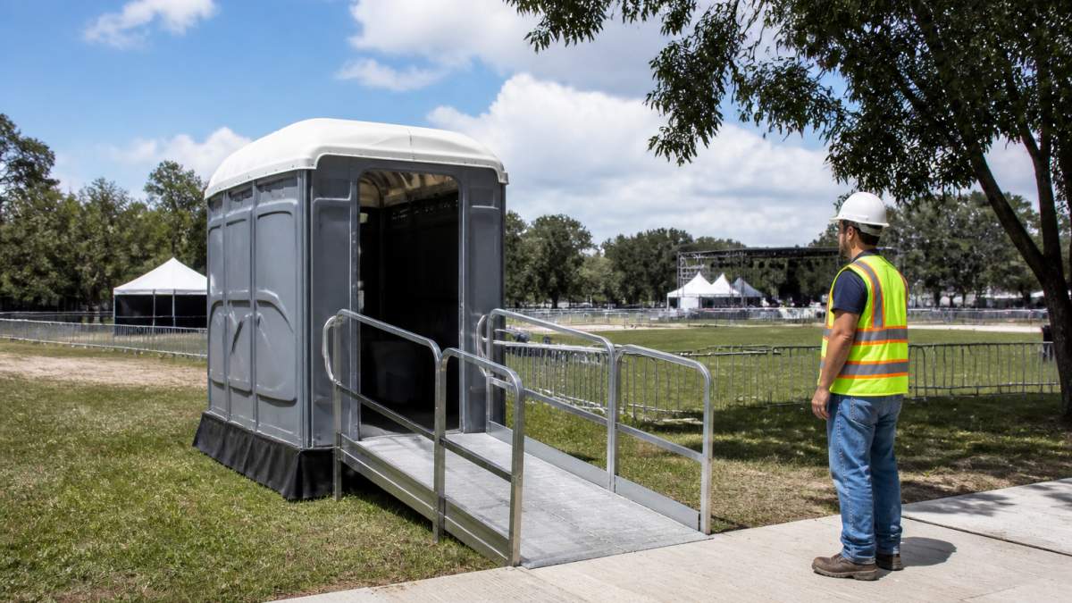 7 ADA Compliant Portable Toilet Rental Options to Compare 1 An ADA-compliant portable toilet with a ramped entrance set on firm level ground at the edge of an outdoor event site, with a visible clear path leading to the door and a site crew member in a hard hat checking placement
