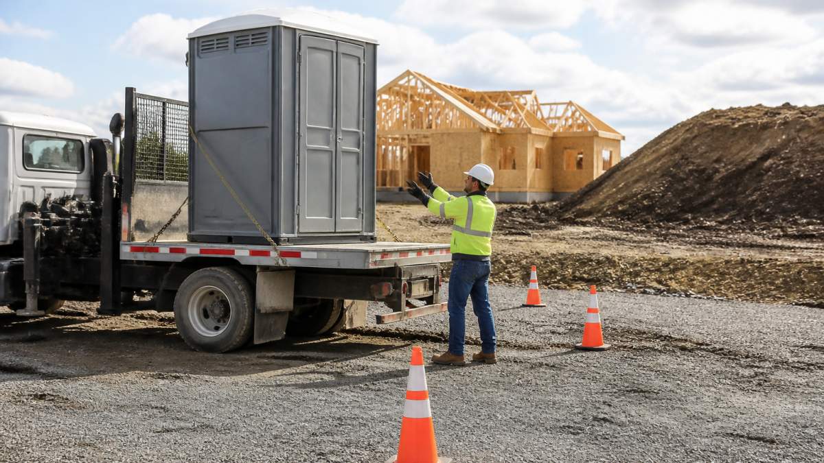 Emergency Same Day Portable Toilet Delivery: What to Expect 1 A portable toilet being delivered by a small truck to a construction site, with a worker guiding the placement on level ground, safety cones visible, and a clear path for the truck