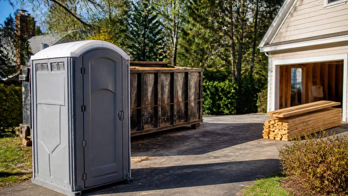 Portable Restroom Rental for Small Renovations: 2026 Guide 1 A standard portable restroom unit placed on a residential driveway beside a home under renovation, with a roll-off dumpster and stacked lumber in the background on a clear afternoon